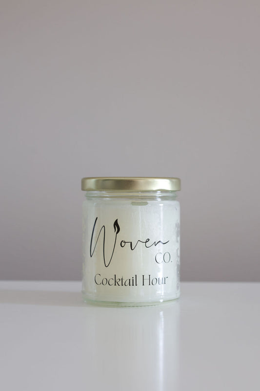 Candle jar labeled 'Woven Candle Co. Cocktail Hour' on a white surface with a light purple background