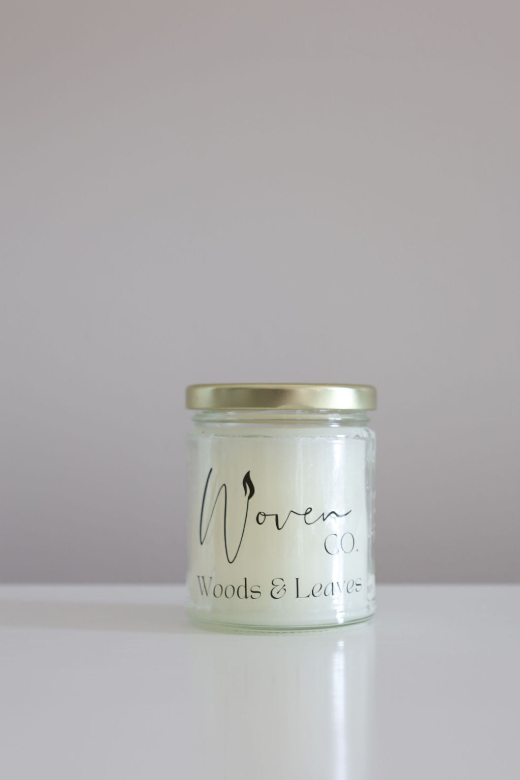 Candle jar labeled 'Woven Co. Woods & Leaves' on a white surface with a light gray background