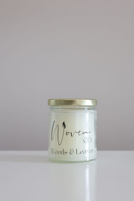 Candle jar labeled 'Woven Co. Woods & Leaves' on a white surface with a light gray background