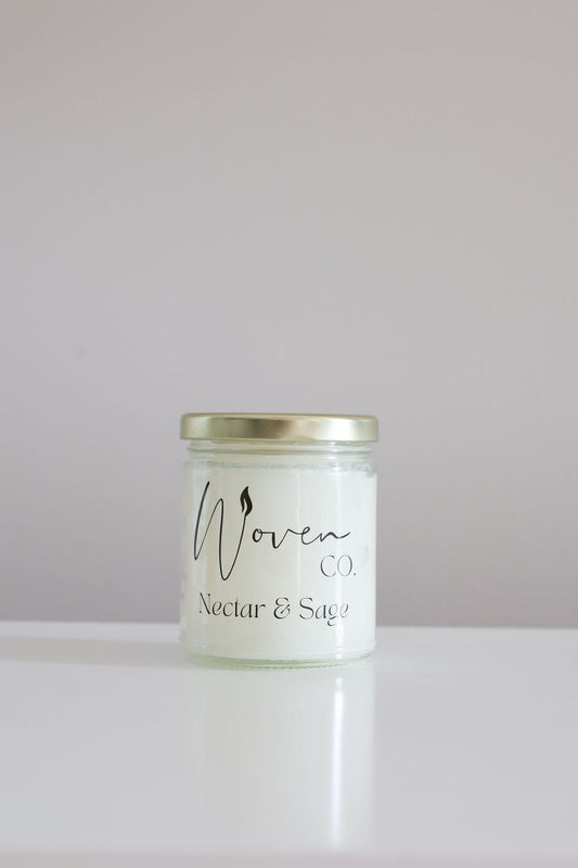 Candle jar labeled 'Woven Co Nectar & Sage' on a white surface with a light gray background