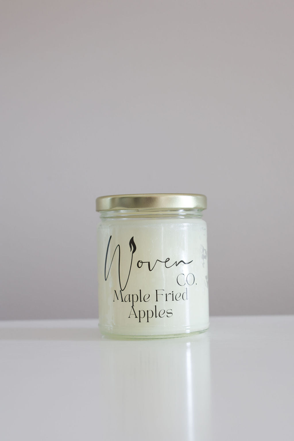 Candle jar labeled 'Maple Fried Apples' by 'Woven Co' on a white surface with a gray background