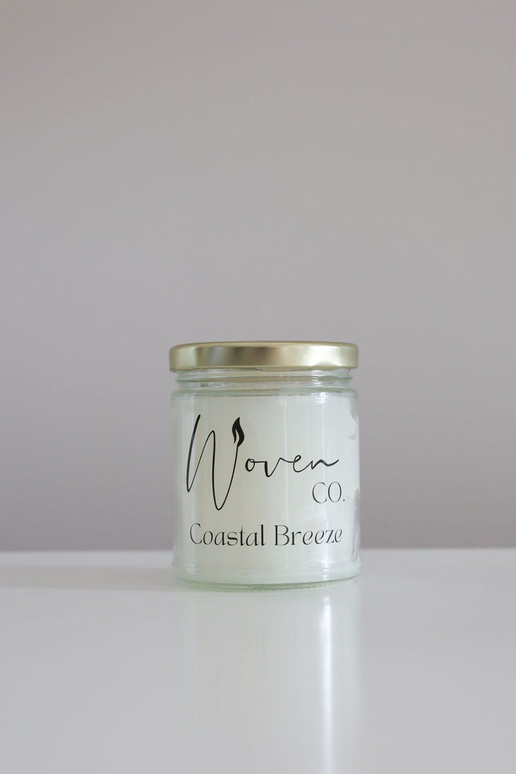 Candle jar labeled 'Woven Coastal Breeze' on a white surface with a light gray background