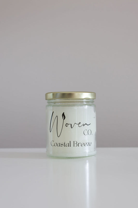 Candle jar labeled 'Woven Coastal Breeze' on a white surface with a light gray background