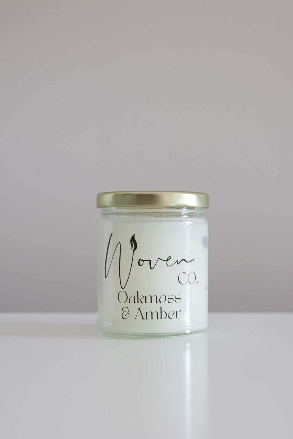Candle jar labeled 'Woven Oakmoss & Amber' on a white background