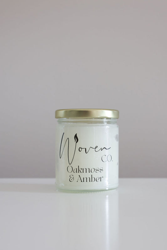Candle jar labeled 'Woven Oakmoss & Amber' on a white background