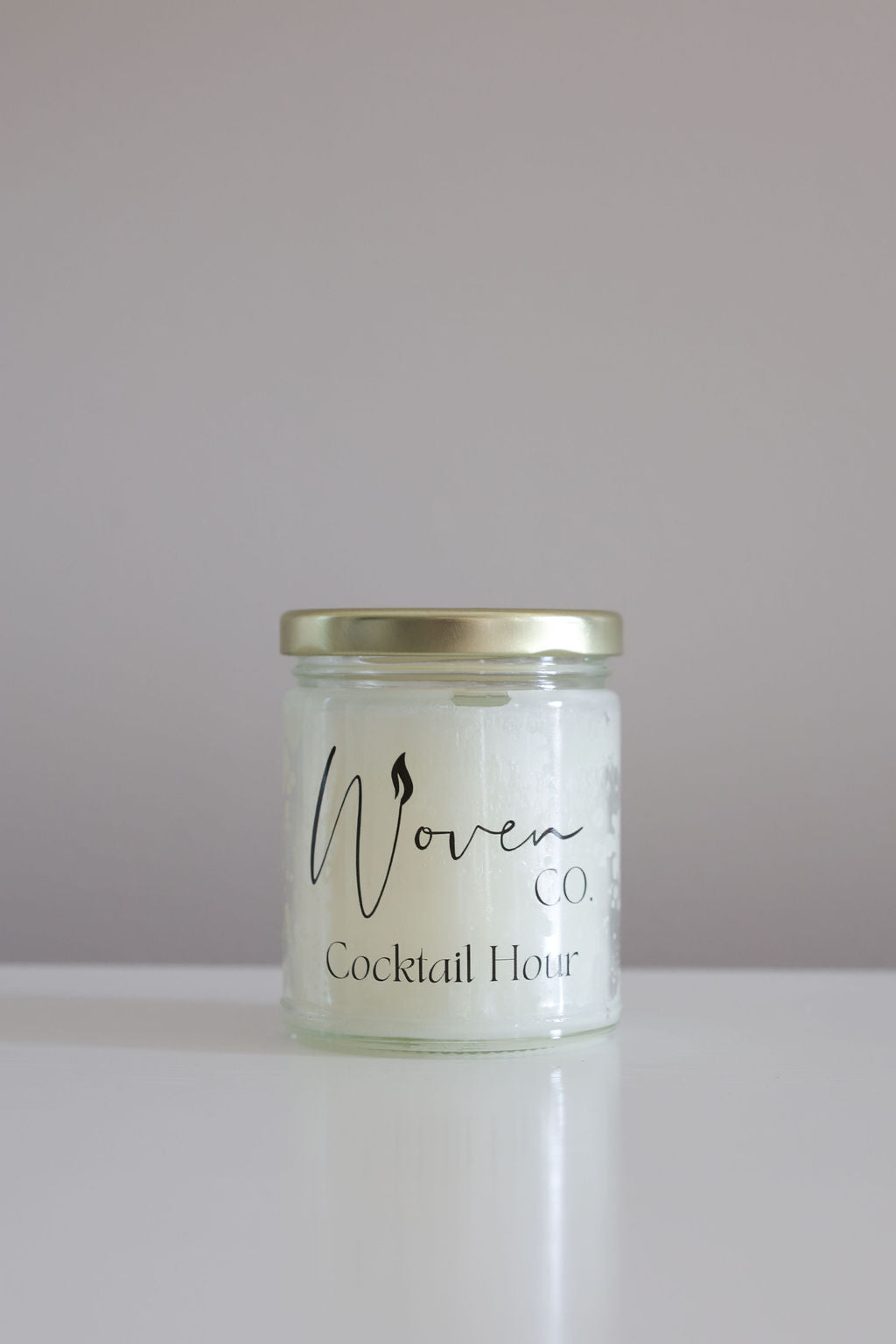 Candle jar labeled 'Woven Candle Co. Cocktail Hour' on a white surface with a light purple background