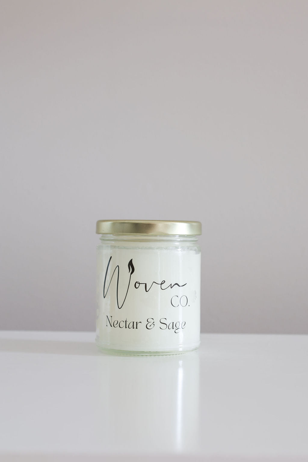 Candle jar labeled 'Woven Co Nectar & Sage' on a white surface with a light gray background