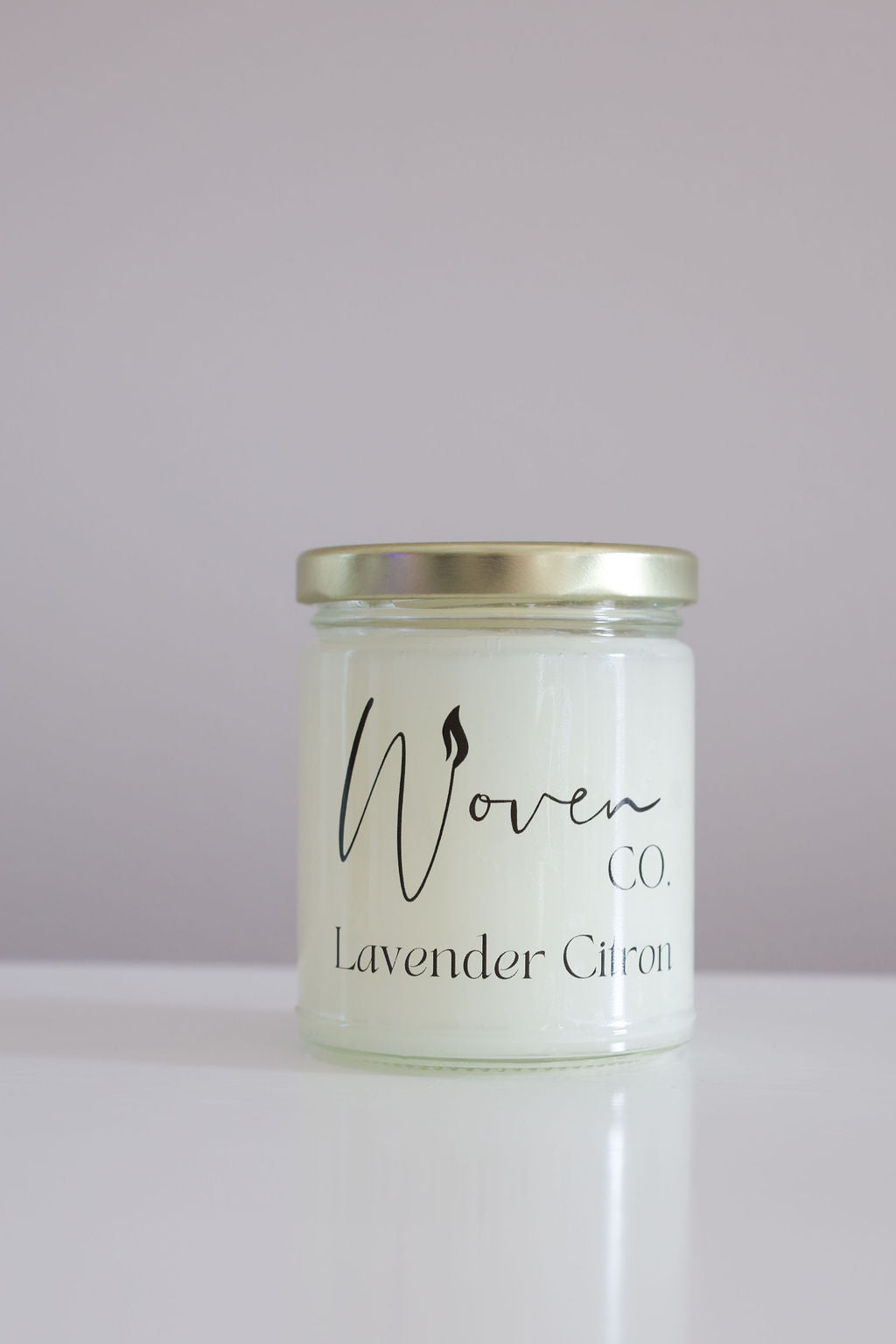 Candle jar labeled 'Woven Co. Lavender Citron' on a white surface with a light purple background